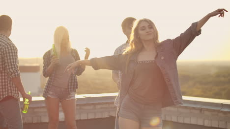 The-young-beautiful-girl-is-dancing-on-the-roof-with-her-four-friends-who-drinks-beer-on-the-party.-She-smiles-and-enjoys-the-time-in-shorts-and-a-light-denim-jacket-in-summer-evening.